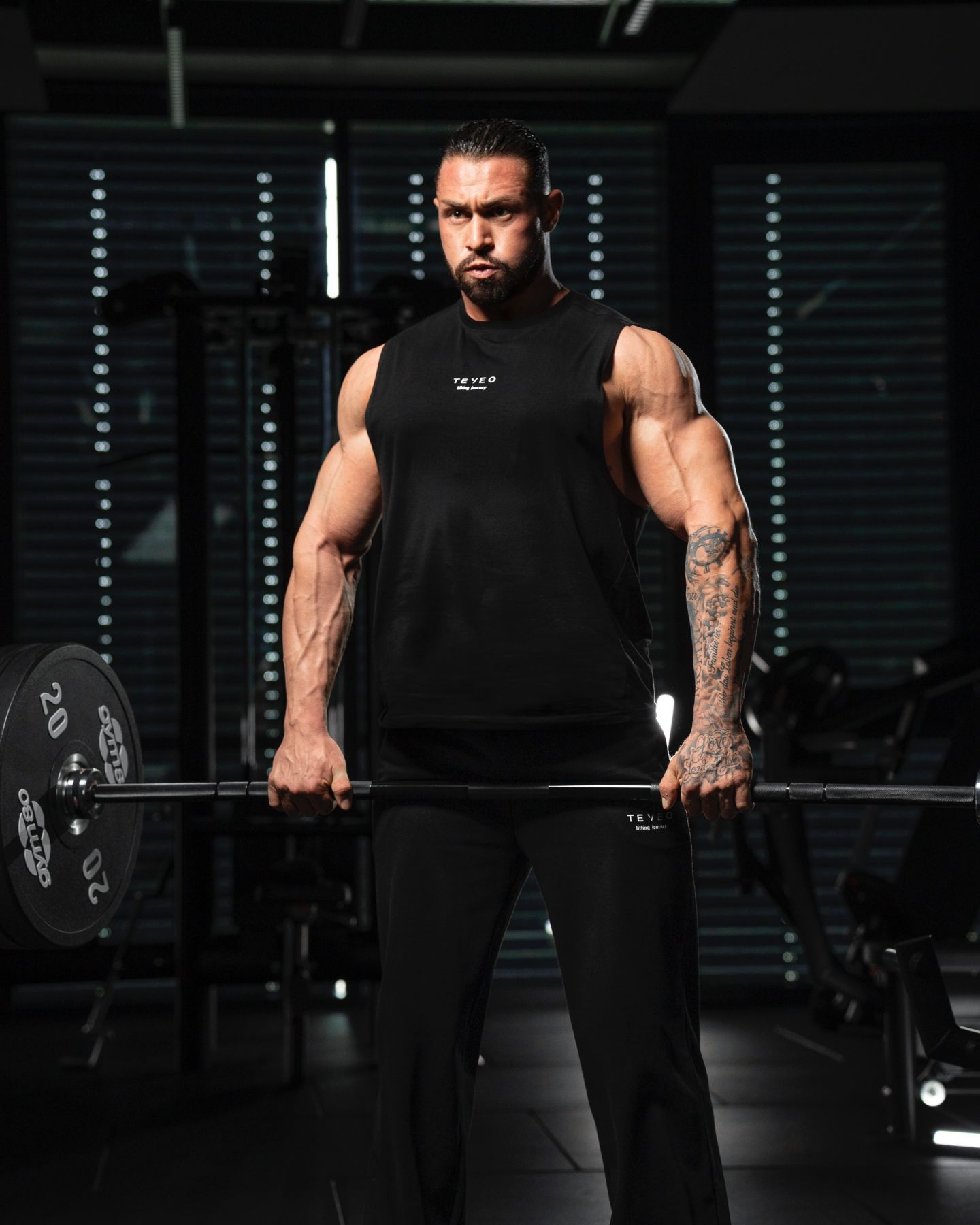 Der Coach in schwarzer Sportkleidung steht im Fitnessstudio und hält eine Langhantel.